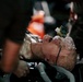 The image depicts what appears to be a medical training mannequin or simulation dummy with realistic wound makeup, surrounded by medical equipment and tubing. The figure has simulated blood and injuries on the head and neck area, consistent with a trauma training scenario. Multiple medical devices, tubes, and personnel hands are visible around the subject.