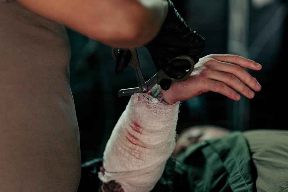A medical or first-aid provider wearing black gloves is using trauma shears (scissors) to cut away bandaging from an injured person's forearm and wrist area. The white gauze bandaging shows visible blood staining, indicating a wound beneath. The patient appears to have tattoos on their arm and is wearing olive/green clothing.