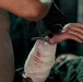 A medical or first-aid provider wearing black gloves is using trauma shears (scissors) to cut away bandaging from an injured person's forearm and wrist area. The white gauze bandaging shows visible blood staining, indicating a wound beneath. The patient appears to have tattoos on their arm and is wearing olive/green clothing.