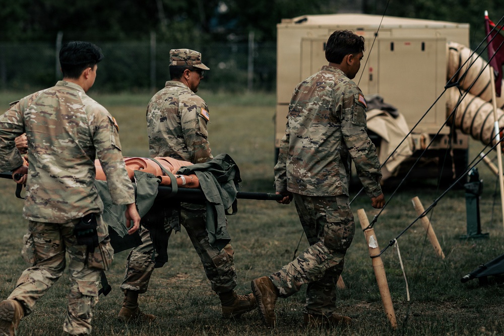 Three soldiers in U.S. Army Operational Camouflage Pattern (OCP) uniforms are carrying an orange litter/stretcher across a grassy field. The scene appears to be a field medical training exercise, indicated by the presence of military field equipment, a tan shelter unit, and tent rigging in the background. All personnel wear American flag patches on their right shoulders.