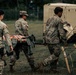 Three soldiers in U.S. Army Operational Camouflage Pattern (OCP) uniforms are carrying an orange litter/stretcher across a grassy field. The scene appears to be a field medical training exercise, indicated by the presence of military field equipment, a tan shelter unit, and tent rigging in the background. All personnel wear American flag patches on their right shoulders.