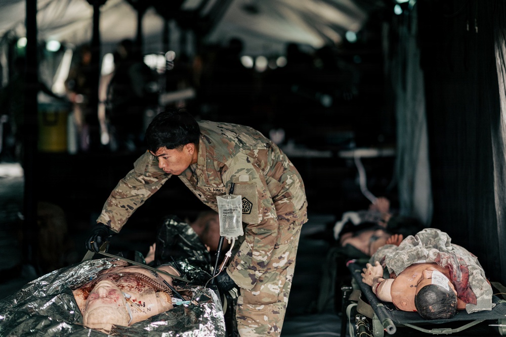 A U.S. Army medic in OCP (Operational Camouflage Pattern) uniform is treating a casualty on a litter inside a military field hospital tent. The medic is administering IV fluids while attending to what appear to be simulated casualties, suggesting this is a medical training exercise. Multiple litters with casualties or mannequins are visible in the background under a large tent structure.