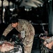 A U.S. Army medic in OCP (Operational Camouflage Pattern) uniform is treating a casualty on a litter inside a military field hospital tent. The medic is administering IV fluids while attending to what appear to be simulated casualties, suggesting this is a medical training exercise. Multiple litters with casualties or mannequins are visible in the background under a large tent structure.