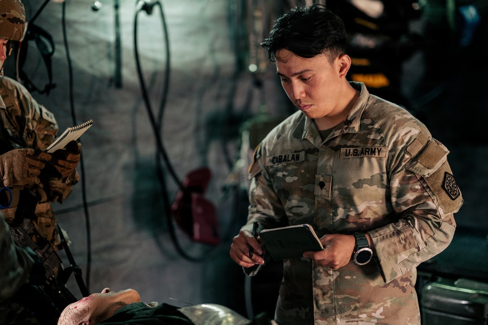 A U.S. Army soldier identified by the name tape 'CABALAN' is taking notes in a small notebook while attending to what appears to be a casualty or training mannequin in a dimly lit enclosed space, possibly a military vehicle or tent. A second soldier in the background is also writing in a notebook. The scene suggests a medical training exercise or field documentation activity.