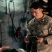 A U.S. Army soldier identified by the name tape 'CABALAN' is taking notes in a small notebook while attending to what appears to be a casualty or training mannequin in a dimly lit enclosed space, possibly a military vehicle or tent. A second soldier in the background is also writing in a notebook. The scene suggests a medical training exercise or field documentation activity.