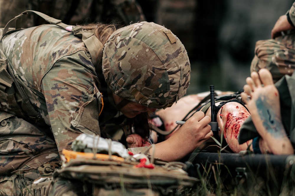 A military medic in MultiCam/OCP camouflage uniform and helmet is providing tactical combat casualty care (TCCC) to a wounded individual on a litter. The medic appears to be writing on or treating a limb that shows visible blood and wounds, with a tourniquet visible on the casualty's leg. This appears to be either a real field medical scenario or a training exercise based on the context.