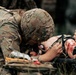A military medic in MultiCam/OCP camouflage uniform and helmet is providing tactical combat casualty care (TCCC) to a wounded individual on a litter. The medic appears to be writing on or treating a limb that shows visible blood and wounds, with a tourniquet visible on the casualty's leg. This appears to be either a real field medical scenario or a training exercise based on the context.
