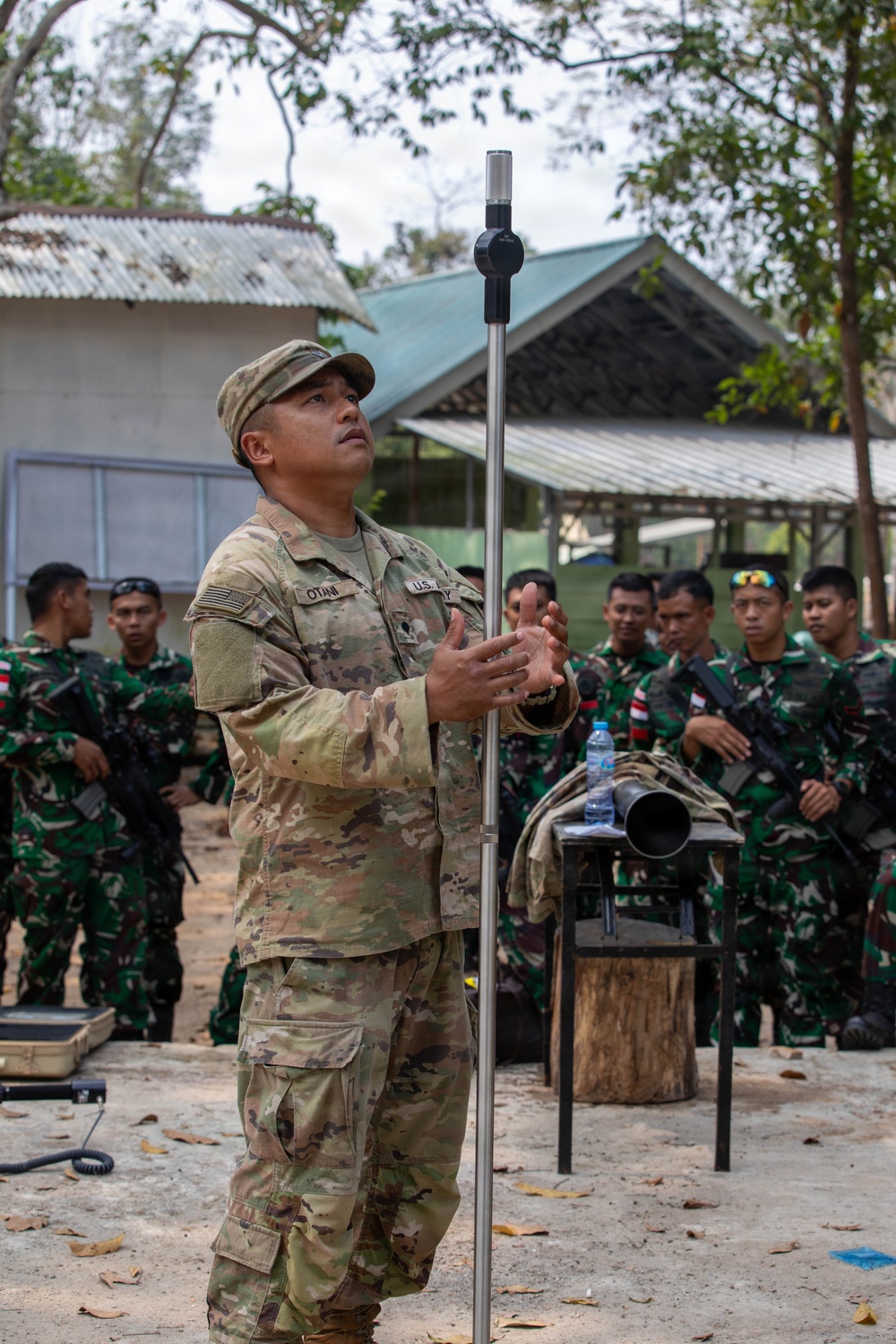 A U.S. Army soldier, identified by name tape as 'OTANI', is conducting a demonstration or instruction session while holding a tall metal rod with a spherical sensor device at the top. He is surrounded by a group of soldiers wearing Indonesian Army (TNI-AD) camouflage uniforms, suggesting a joint training or exchange exercise. The setting appears to be an outdoor training area with basic structures in the background.