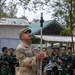 A U.S. Army soldier, identified by name tape as 'OTANI', is conducting a demonstration or instruction session while holding a tall metal rod with a spherical sensor device at the top. He is surrounded by a group of soldiers wearing Indonesian Army (TNI-AD) camouflage uniforms, suggesting a joint training or exchange exercise. The setting appears to be an outdoor training area with basic structures in the background.