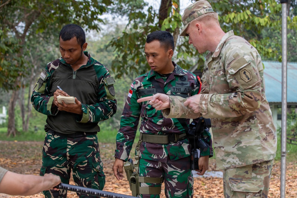 Three military personnel are engaged in what appears to be a training or briefing session in an outdoor wooded area. Two soldiers wearing Indonesian Army (TNI AD) camouflage uniforms are interacting with a U.S. Army soldier in MultiCam/OCP pattern uniform, who is pointing and providing instruction. One Indonesian soldier is taking notes while the other listens attentively.