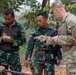 Three military personnel are engaged in what appears to be a training or briefing session in an outdoor wooded area. Two soldiers wearing Indonesian Army (TNI AD) camouflage uniforms are interacting with a U.S. Army soldier in MultiCam/OCP pattern uniform, who is pointing and providing instruction. One Indonesian soldier is taking notes while the other listens attentively.
