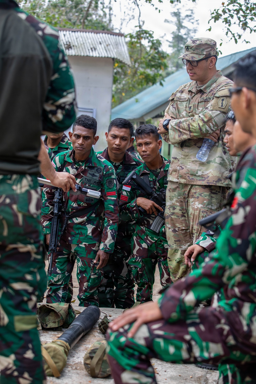 A U.S. Army soldier in OCP (Operational Camouflage Pattern) uniform observes Indonesian Army (TNI AD) soldiers during what appears to be a joint military training exercise. The Indonesian soldiers are wearing their distinctive green woodland camouflage uniforms with Indonesian flag patches and are gathered around what appears to be a weapons or equipment demonstration. The setting is an outdoor military training area with structures visible in the background.