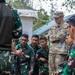 A U.S. Army soldier in OCP (Operational Camouflage Pattern) uniform observes Indonesian Army (TNI AD) soldiers during what appears to be a joint military training exercise. The Indonesian soldiers are wearing their distinctive green woodland camouflage uniforms with Indonesian flag patches and are gathered around what appears to be a weapons or equipment demonstration. The setting is an outdoor military training area with structures visible in the background.