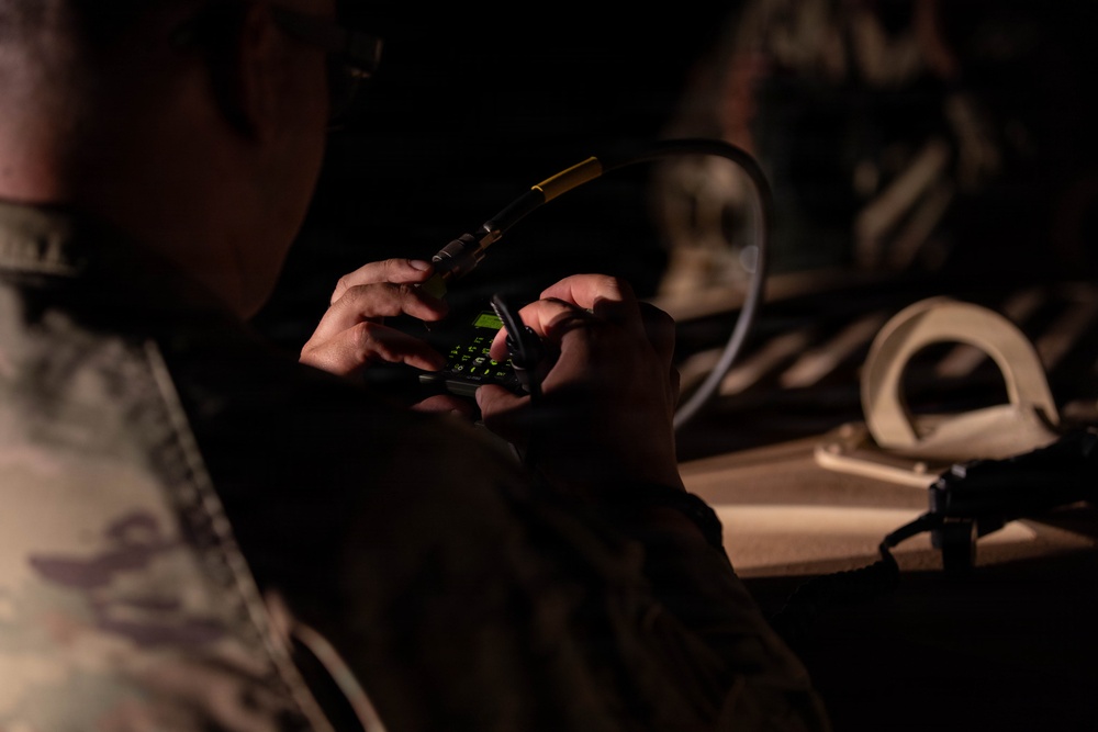 A military personnel wearing camouflage uniform is operating a handheld communications device or radio in a low-light environment. The individual appears to be inputting data or programming the device, which displays a green-lit keypad. The scene suggests a tactical or field operations setting.