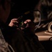 A military personnel wearing camouflage uniform is operating a handheld communications device or radio in a low-light environment. The individual appears to be inputting data or programming the device, which displays a green-lit keypad. The scene suggests a tactical or field operations setting.