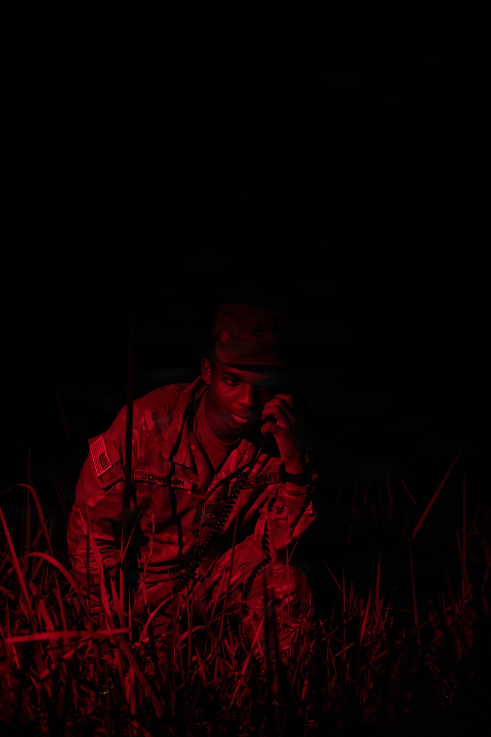 A U.S. Army soldier is photographed at night in a field, illuminated by red light. The soldier is crouching or sitting low in tall grass while holding a radio handset to their ear. The name tape on the uniform reads 'JOHNSON' and a U.S. flag patch is visible on the left shoulder.