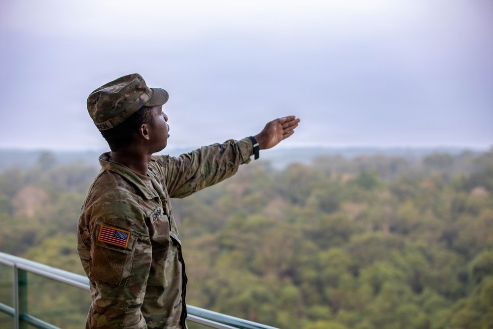 A U.S. Army soldier in Operational Camouflage Pattern (OCP) uniform is standing on an elevated platform or observation deck, extending one arm outward in a pointing or directing gesture. The soldier is wearing a patrol cap and has a U.S. flag patch visible on the left sleeve. A forested landscape stretches into the background under an overcast sky.