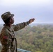 A U.S. Army soldier in Operational Camouflage Pattern (OCP) uniform is standing on an elevated platform or observation deck, extending one arm outward in a pointing or directing gesture. The soldier is wearing a patrol cap and has a U.S. flag patch visible on the left sleeve. A forested landscape stretches into the background under an overcast sky.