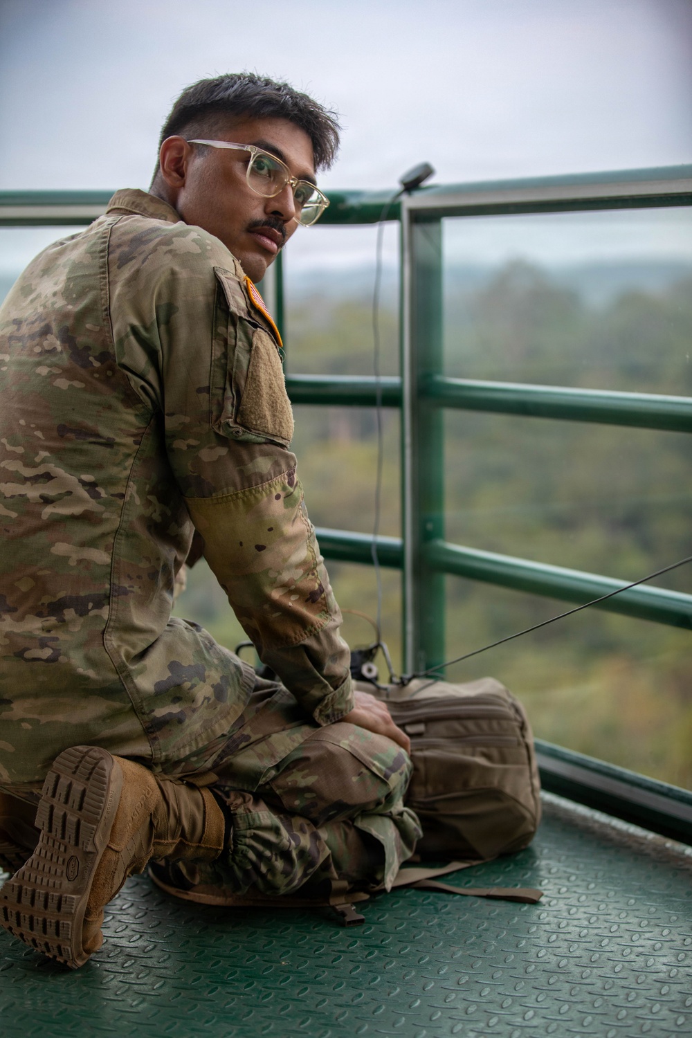A young male soldier in US Army OCP (Operational Camouflage Pattern) uniform is seated cross-legged on an elevated green metal platform, looking back over his shoulder toward the camera. He is positioned near a green safety railing with a wooded, hilly landscape visible in the background under an overcast sky. A tactical bag or pouch rests in his lap, and a cable or wire is visible near the railing.