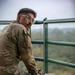 A young male soldier in US Army OCP (Operational Camouflage Pattern) uniform is seated cross-legged on an elevated green metal platform, looking back over his shoulder toward the camera. He is positioned near a green safety railing with a wooded, hilly landscape visible in the background under an overcast sky. A tactical bag or pouch rests in his lap, and a cable or wire is visible near the railing.