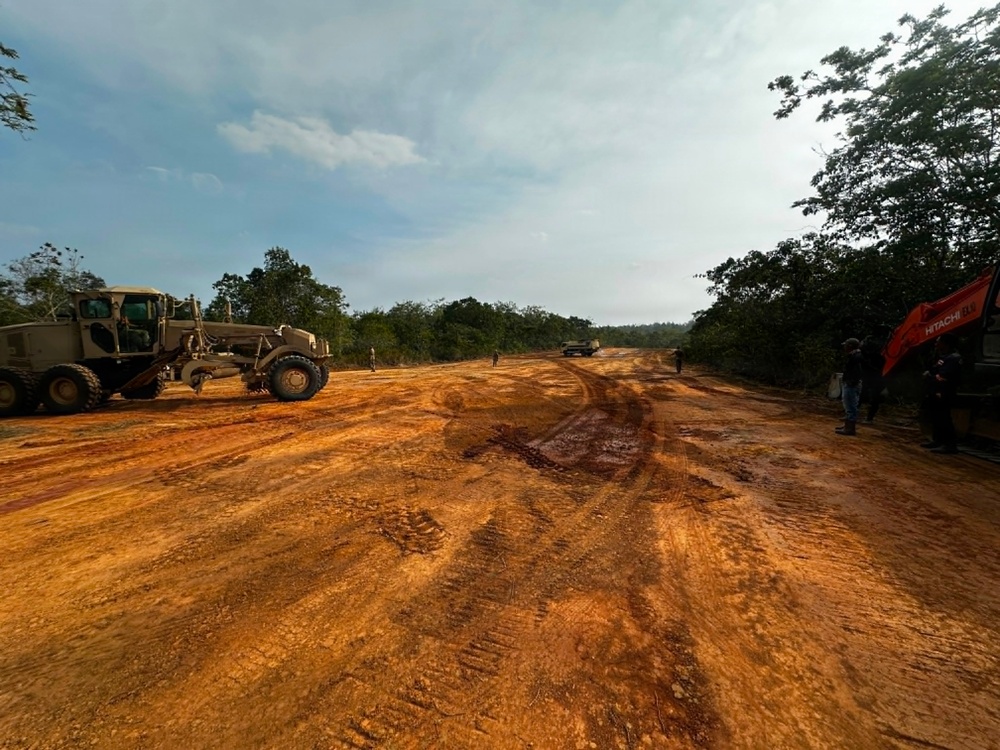 The image shows an active road construction or clearing operation in a forested area, featuring heavy machinery working on a wide dirt track with red-orange laterite soil. Multiple pieces of construction equipment are visible alongside several workers on what appears to be a newly graded road corridor cut through tropical vegetation. The scene suggests ongoing infrastructure development in a rural or remote forested region.