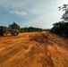 The image shows an active road construction or clearing operation in a forested area, featuring heavy machinery working on a wide dirt track with red-orange laterite soil. Multiple pieces of construction equipment are visible alongside several workers on what appears to be a newly graded road corridor cut through tropical vegetation. The scene suggests ongoing infrastructure development in a rural or remote forested region.