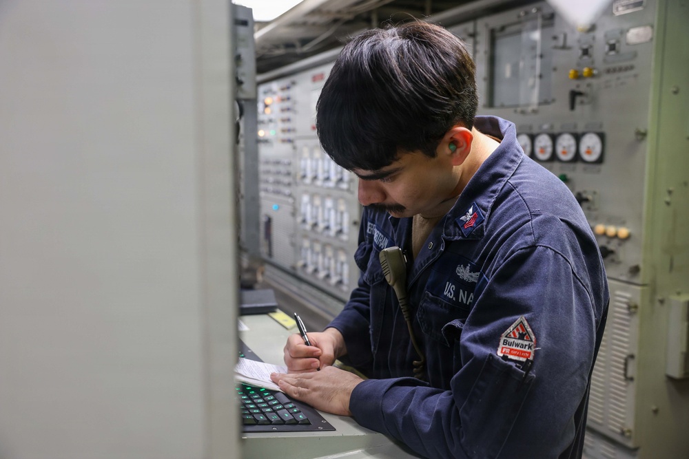 A U.S. Navy sailor in a dark blue coverall uniform is writing notes on a document while standing at a control panel aboard a naval vessel. The individual appears to be conducting an inspection, logging data, or performing a routine operational task in what appears to be an engineering or combat systems space. The environment is filled with switches, gauges, and control panels typical of a naval ship's interior.