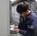 A U.S. Navy sailor in a dark blue coverall uniform is writing notes on a document while standing at a control panel aboard a naval vessel. The individual appears to be conducting an inspection, logging data, or performing a routine operational task in what appears to be an engineering or combat systems space. The environment is filled with switches, gauges, and control panels typical of a naval ship's interior.