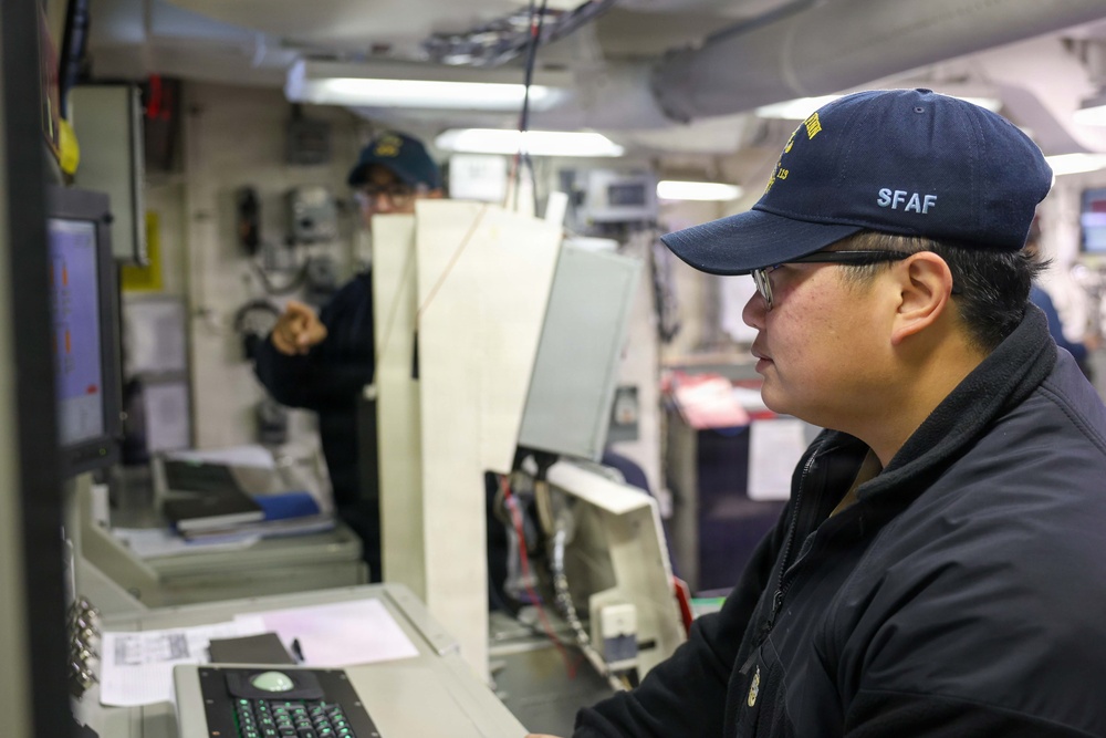 Two personnel are working inside what appears to be a naval vessel's operations or combat information center. The foreground individual, wearing a navy blue cap marked 'SFAF' and glasses, is focused on a monitor and control console. A second crew member is visible in the background, appearing to gesture or point at something.