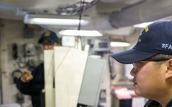 Two personnel are working inside what appears to be a naval vessel's operations or combat information center. The foreground individual, wearing a navy blue cap marked 'SFAF' and glasses, is focused on a monitor and control console. A second crew member is visible in the background, appearing to gesture or point at something.