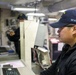 Two personnel are working inside what appears to be a naval vessel's operations or combat information center. The foreground individual, wearing a navy blue cap marked 'SFAF' and glasses, is focused on a monitor and control console. A second crew member is visible in the background, appearing to gesture or point at something.