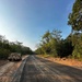 A newly graded dirt/gravel road cuts through a tropical forest setting under a clear blue sky. A military-style HMMWV (Humvee) vehicle is parked on the left shoulder of the road. The road surface shows fresh compaction with visible tire tracks and reddish-orange laterite soil on the shoulders, suggesting recent construction or maintenance.