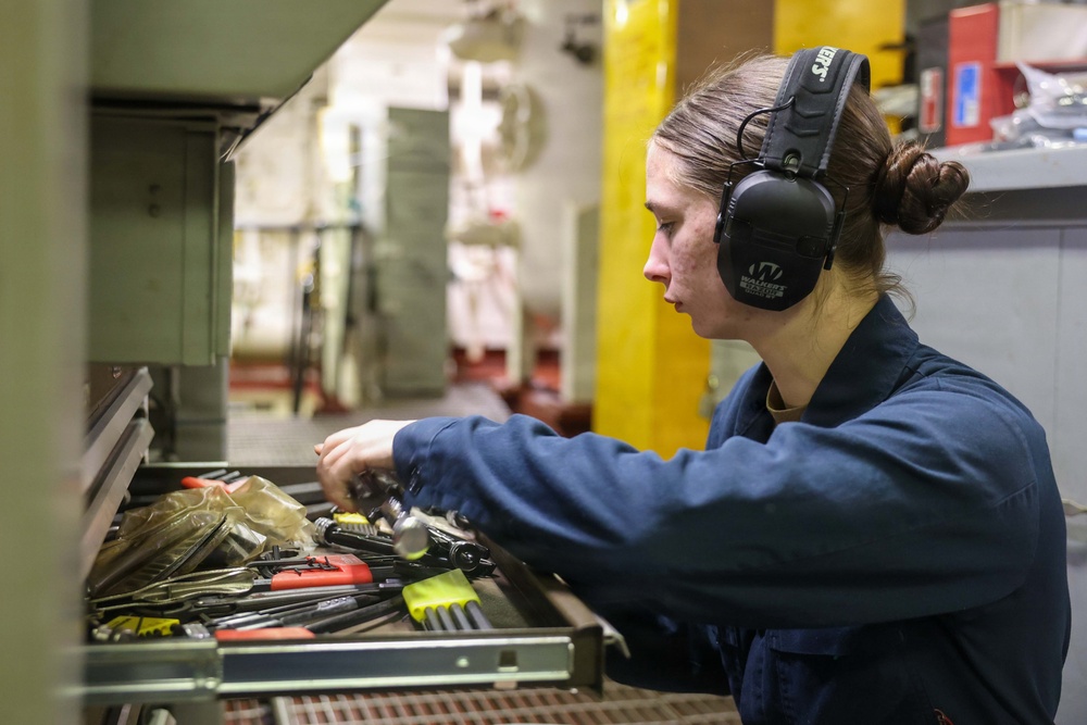 A young woman wearing navy blue coveralls and black Walker's brand hearing protection earmuffs is searching through an open tool drawer in what appears to be an industrial or military maintenance workspace. She has her hair pulled back in a bun and is focused on selecting a tool from a well-stocked drawer containing various hand tools. The background shows a larger workshop or hangar-like environment with yellow structural columns.