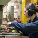 A young woman wearing navy blue coveralls and black Walker's brand hearing protection earmuffs is searching through an open tool drawer in what appears to be an industrial or military maintenance workspace. She has her hair pulled back in a bun and is focused on selecting a tool from a well-stocked drawer containing various hand tools. The background shows a larger workshop or hangar-like environment with yellow structural columns.