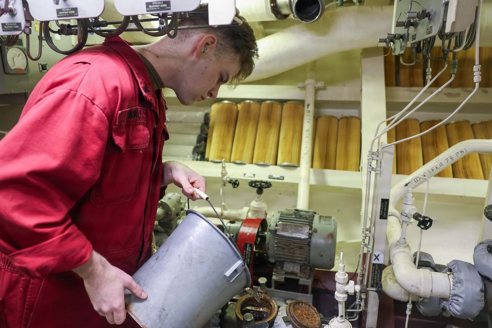 A technician in a red coverall uniform is working in what appears to be a ship's engine room or mechanical space. He is holding a metal bucket and using a dipstick or probe to check fluid levels or take a sample. The background shows industrial equipment including filters, piping, valves, gauges, and an electric motor.