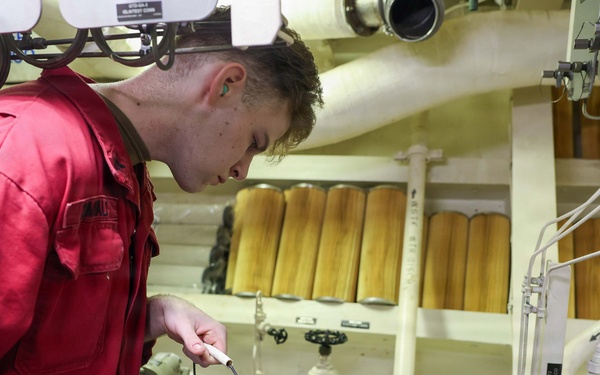 A technician wearing a red coverall uniform is working in what appears to be a ship's engine room or mechanical space. He is holding a metal bucket and using a dipstick or probe to check fluid levels in a container below. The space is filled with industrial equipment including filters, pipes, valves, and an electric motor.
