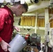 A technician in a red coverall uniform is working in what appears to be a ship's engine room or mechanical space. He is holding a metal bucket and using a dipstick or probe to check fluid levels or take a sample. The background shows industrial equipment including filters, piping, valves, gauges, and an electric motor.