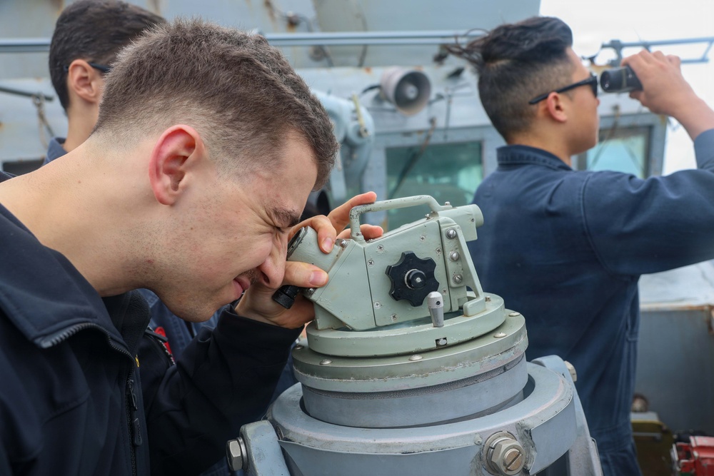 Three individuals in dark navy-style uniforms are on the deck of a military vessel performing navigation and observation duties. The foreground figure is looking through a pelorus or bearing circle mounted on a pedestal, while a second person uses binoculars in the background. A third person is partially visible at the far left.