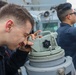 Three individuals in dark navy-style uniforms are on the deck of a military vessel performing navigation and observation duties. The foreground figure is looking through a pelorus or bearing circle mounted on a pedestal, while a second person uses binoculars in the background. A third person is partially visible at the far left.