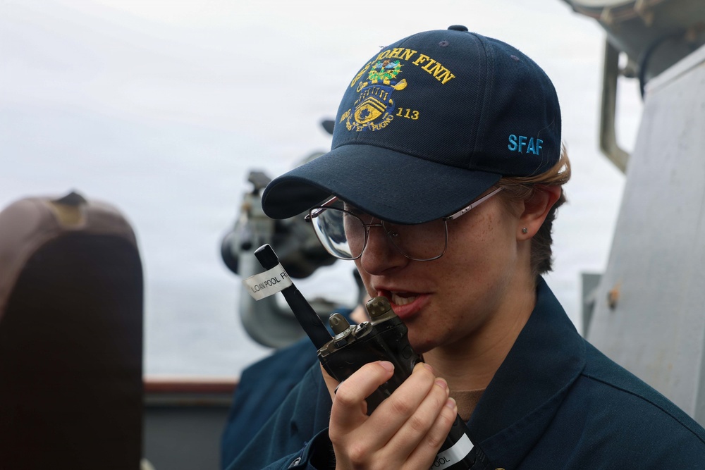 A sailor aboard a U.S. Navy vessel is speaking into a handheld radio/walkie-talkie. They are wearing a navy blue cap embroidered with 'USS JOHN FINN 113' and 'SFAF', along with a matching navy blue uniform. The setting appears to be the bridge or exterior deck of a naval destroyer.