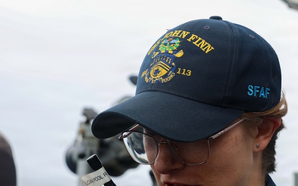 A sailor aboard a U.S. Navy vessel is speaking into a handheld radio/walkie-talkie. They are wearing a navy blue cap embroidered with 'USS JOHN FINN 113' and 'SFAF', along with a matching navy blue uniform. The setting appears to be the exterior deck or bridge wing of a naval destroyer.