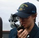 A sailor aboard a U.S. Navy vessel is speaking into a handheld radio/walkie-talkie. They are wearing a navy blue cap embroidered with 'USS JOHN FINN 113' and 'SFAF', along with a matching navy blue uniform. The setting appears to be the bridge or exterior deck of a naval destroyer.