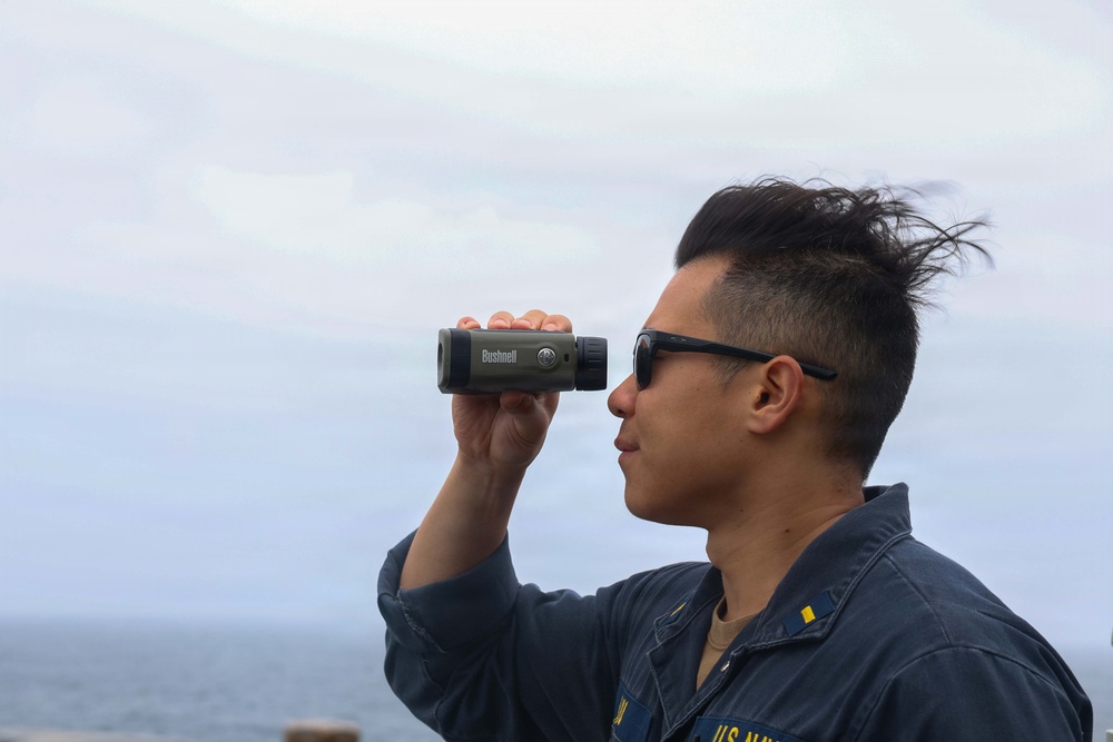 A U.S. Navy service member is using a Bushnell monocular/rangefinder while standing outdoors near a body of water. The individual is wearing a navy blue uniform with 'U.S. Navy' visible on the chest and sunglasses pushed up on their face. The background shows an overcast sky and what appears to be an ocean or large body of water.