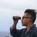 A U.S. Navy service member is using a Bushnell monocular/rangefinder while standing outdoors near a body of water. The individual is wearing a navy blue uniform with 'U.S. Navy' visible on the chest and sunglasses pushed up on their face. The background shows an overcast sky and what appears to be an ocean or large body of water.