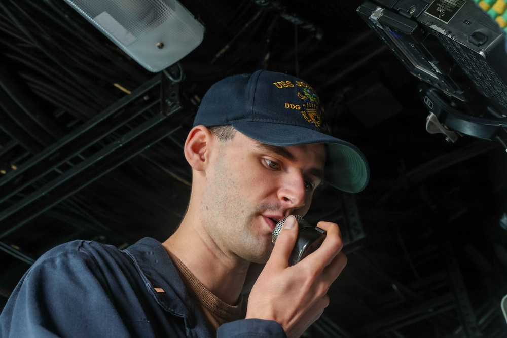 A U.S. Navy sailor is speaking into a radio handset or sound-powered telephone aboard a naval vessel. He is wearing a navy blue ball cap embroidered with 'USS' and 'DDG' designations, indicating assignment to a destroyer-class ship. The image is taken from a low angle in what appears to be an interior shipboard space with overhead equipment and cabling visible.