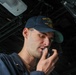 A U.S. Navy sailor is speaking into a radio handset or sound-powered telephone aboard a naval vessel. He is wearing a navy blue ball cap embroidered with 'USS' and 'DDG' designations, indicating assignment to a destroyer-class ship. The image is taken from a low angle in what appears to be an interior shipboard space with overhead equipment and cabling visible.