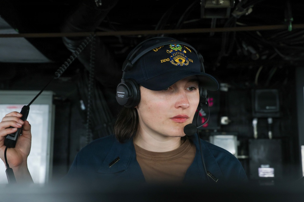 A person wearing a U.S. Navy ball cap marked 'DDG' (destroyer designation) and a Sennheiser communications headset with microphone is holding a coiled cable or handset. The setting appears to be the bridge or interior of a naval vessel, with visible wiring, cables, and equipment panels in the background. The individual appears focused and engaged in an operational or communications task.