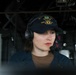 A person wearing a U.S. Navy ball cap marked 'DDG' (destroyer designation) and a Sennheiser communications headset with microphone is holding a coiled cable or handset. The setting appears to be the bridge or interior of a naval vessel, with visible wiring, cables, and equipment panels in the background. The individual appears focused and engaged in an operational or communications task.