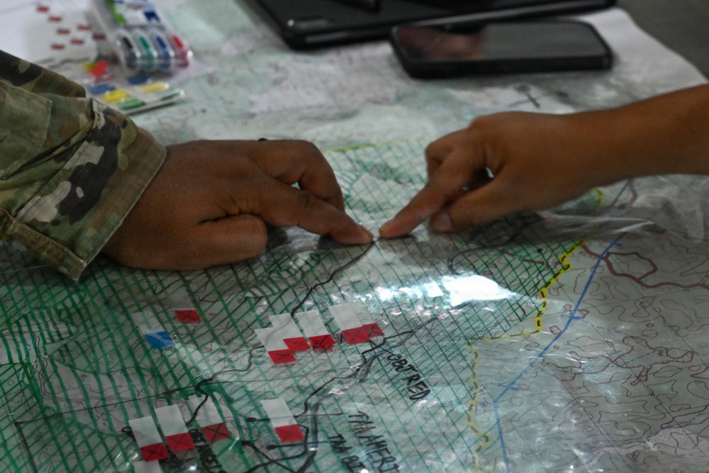 Two individuals are collaborating over a military tactical map covered with a clear acetate overlay, marked with colored symbols and grid lines. One person is wearing a camouflage uniform, indicating military personnel. They appear to be conducting a map briefing or operational planning session, pointing to specific locations on the map.