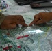 Two individuals are collaborating over a military tactical map covered with a clear acetate overlay, marked with colored symbols and grid lines. One person is wearing a camouflage uniform, indicating military personnel. They appear to be conducting a map briefing or operational planning session, pointing to specific locations on the map.